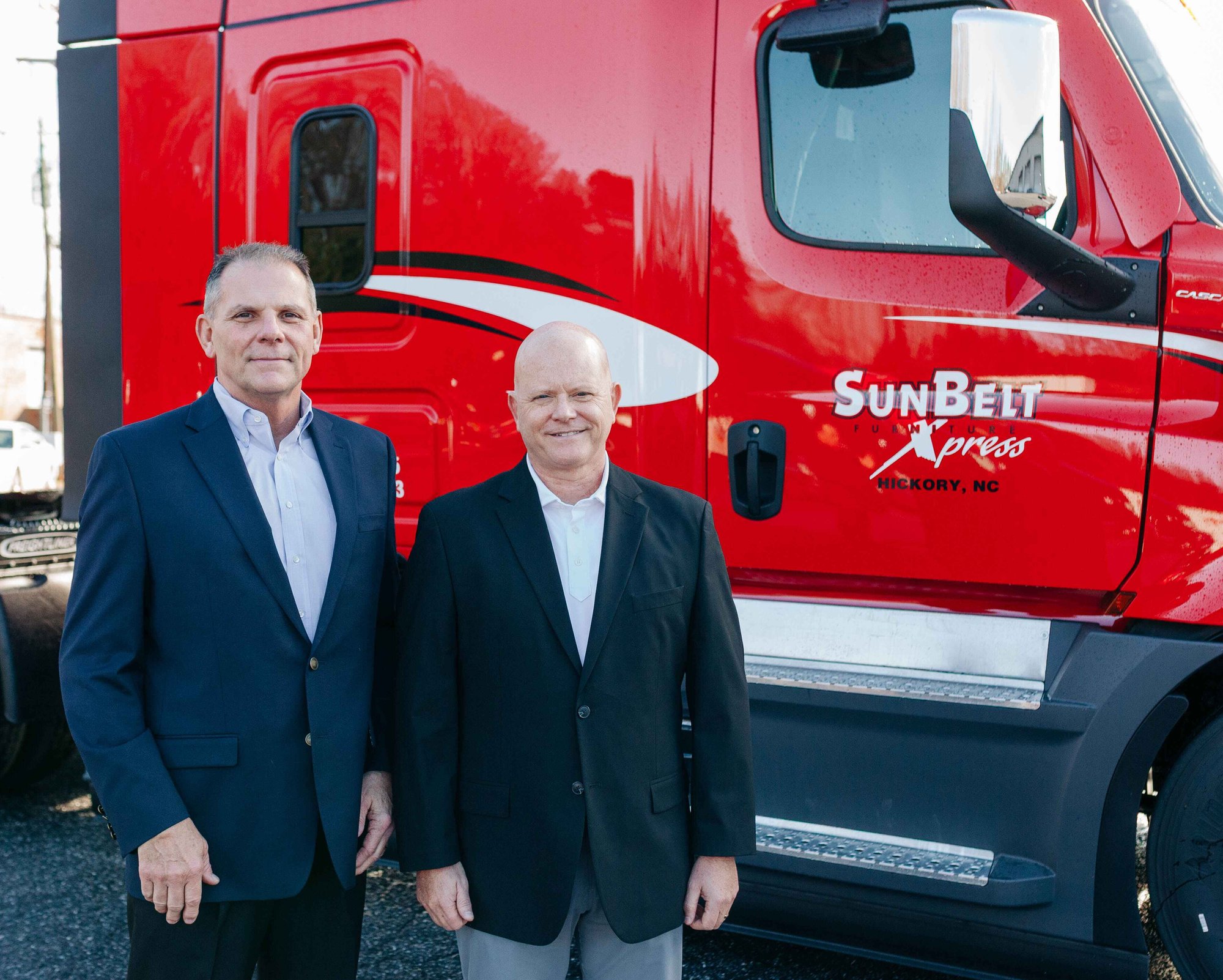 Two men stand in front of a bright red semi truck, smiling and posing for the photo.