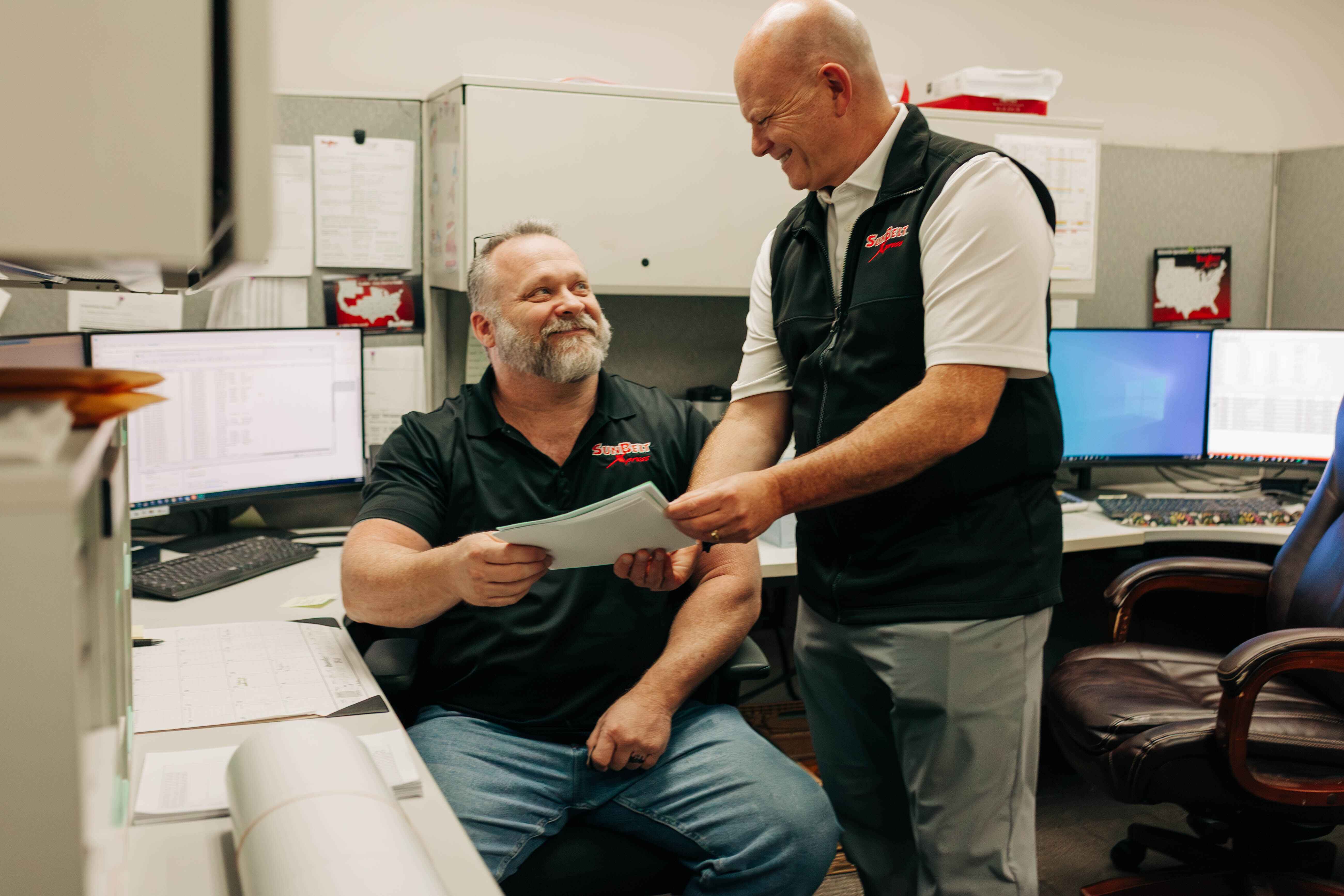 Two men in an office discussing and holding papers, looking engaged in conversation.